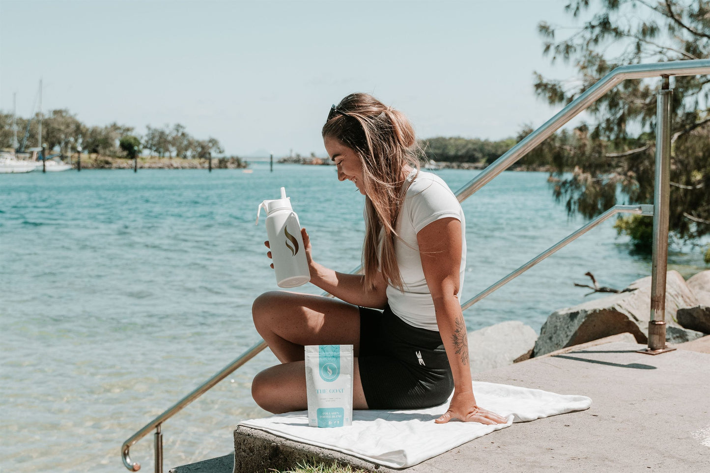 Woman laughing by turquoise water with a Brewdy bottle and The GOAT collagen coffee